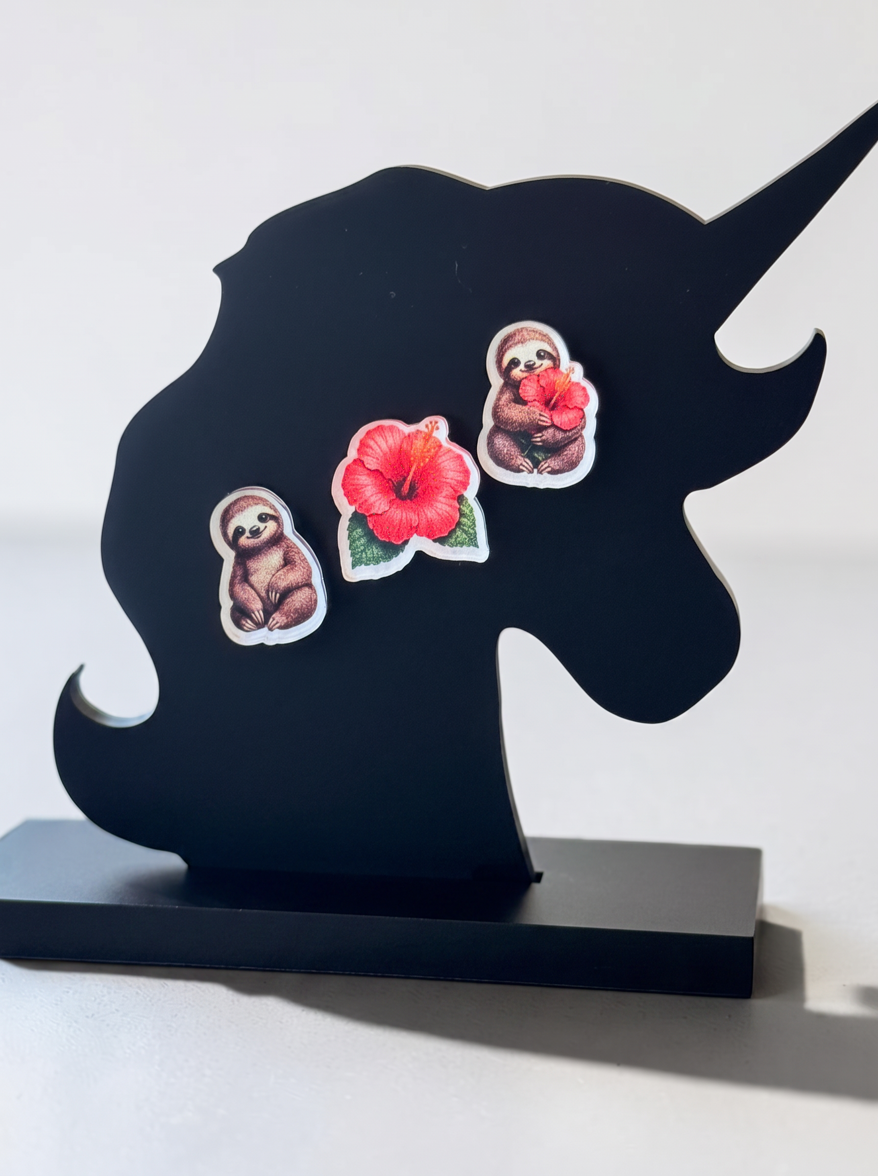 A black unicorn-shaped decorative plaque or stand with a rectangular base, featuring three small decorative earrings: two sloths and a hibiscus flower, displayed against a plain light background.