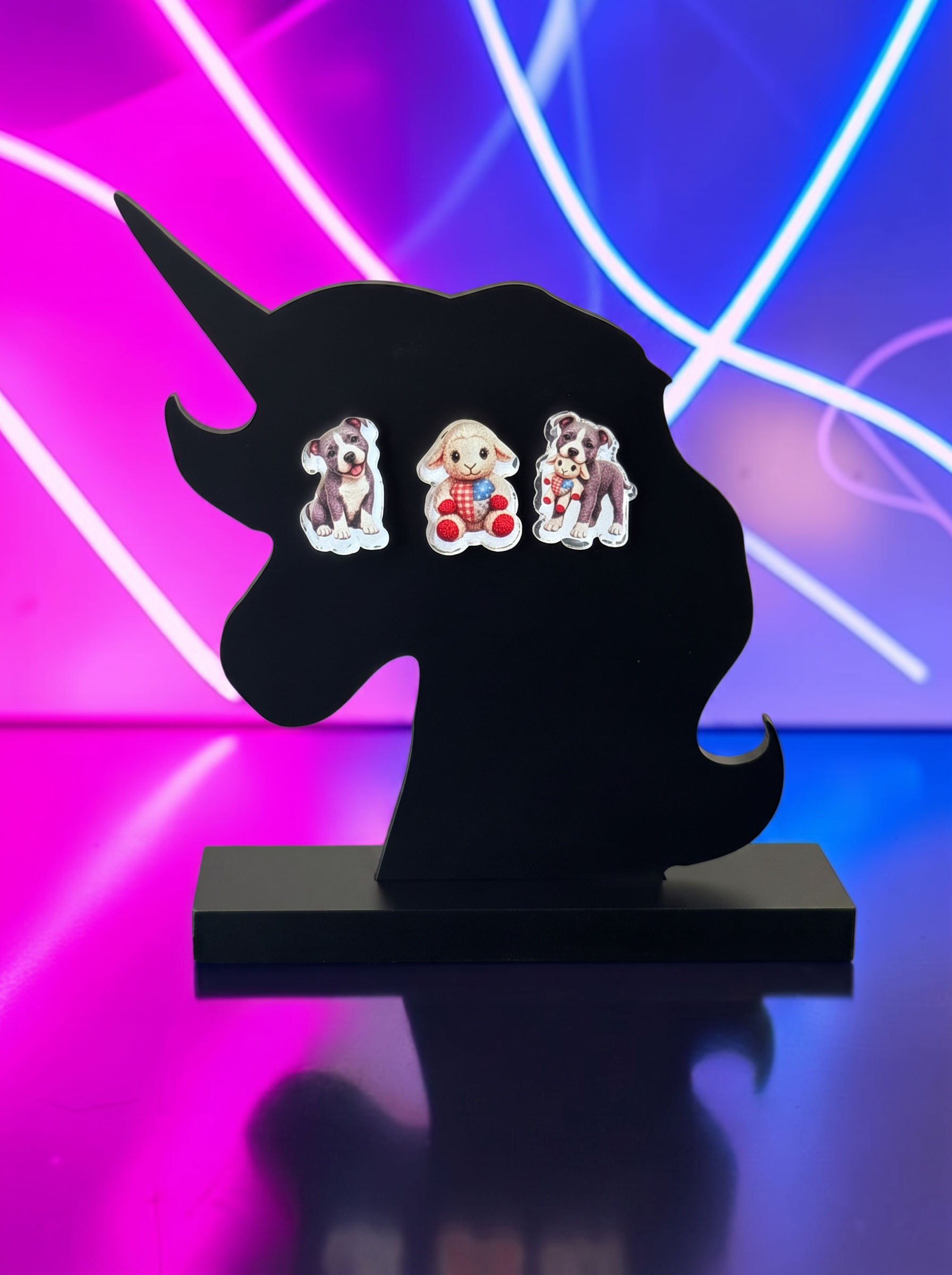 Three small pitt bull themed acrylic earrings displayed on a black unicorn-shaped stand against a vibrant pink and blue neon light background.