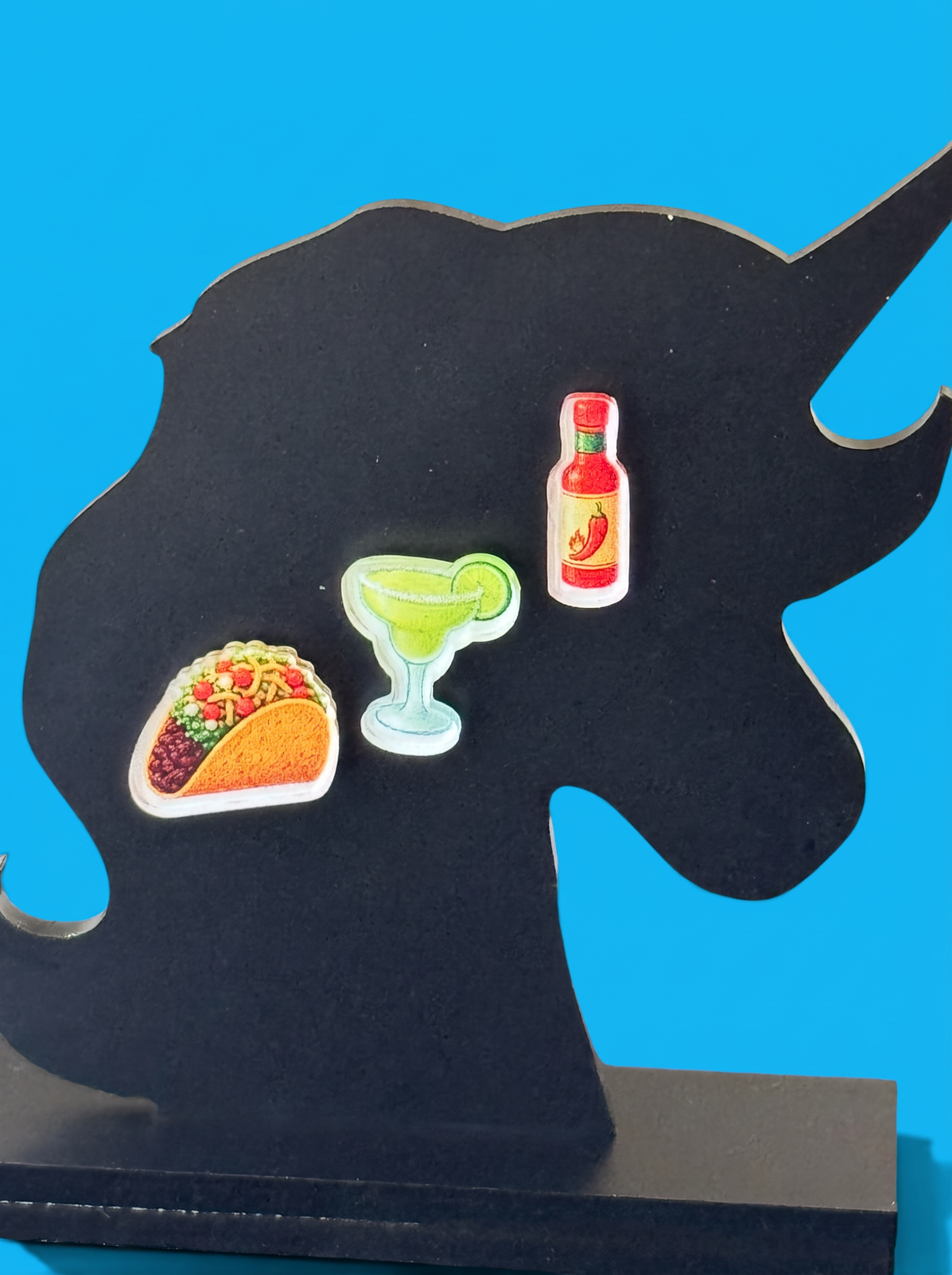 Three small novelty earrings shaped like a taco, a margarita glass, and a hot sauce bottle, displayed on a black unicorn silhouette stand against a bright blue background.