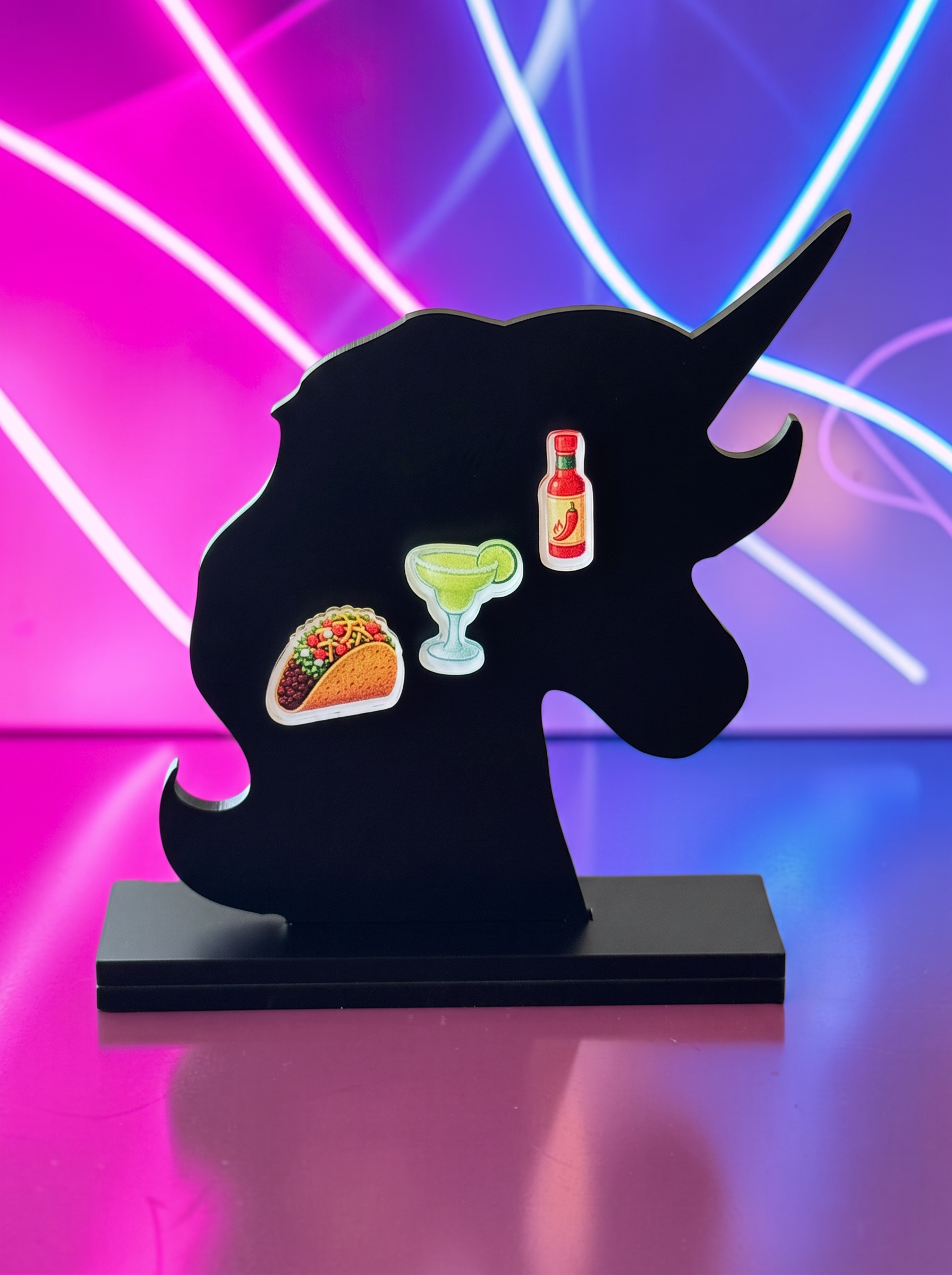A black unicorn-shaped decorative plaque on a matching rectangular base, featuring small colorful earrings of a taco, a margarita glass, and a hot sauce bottle, set against a vibrant pink and purple neon light background.