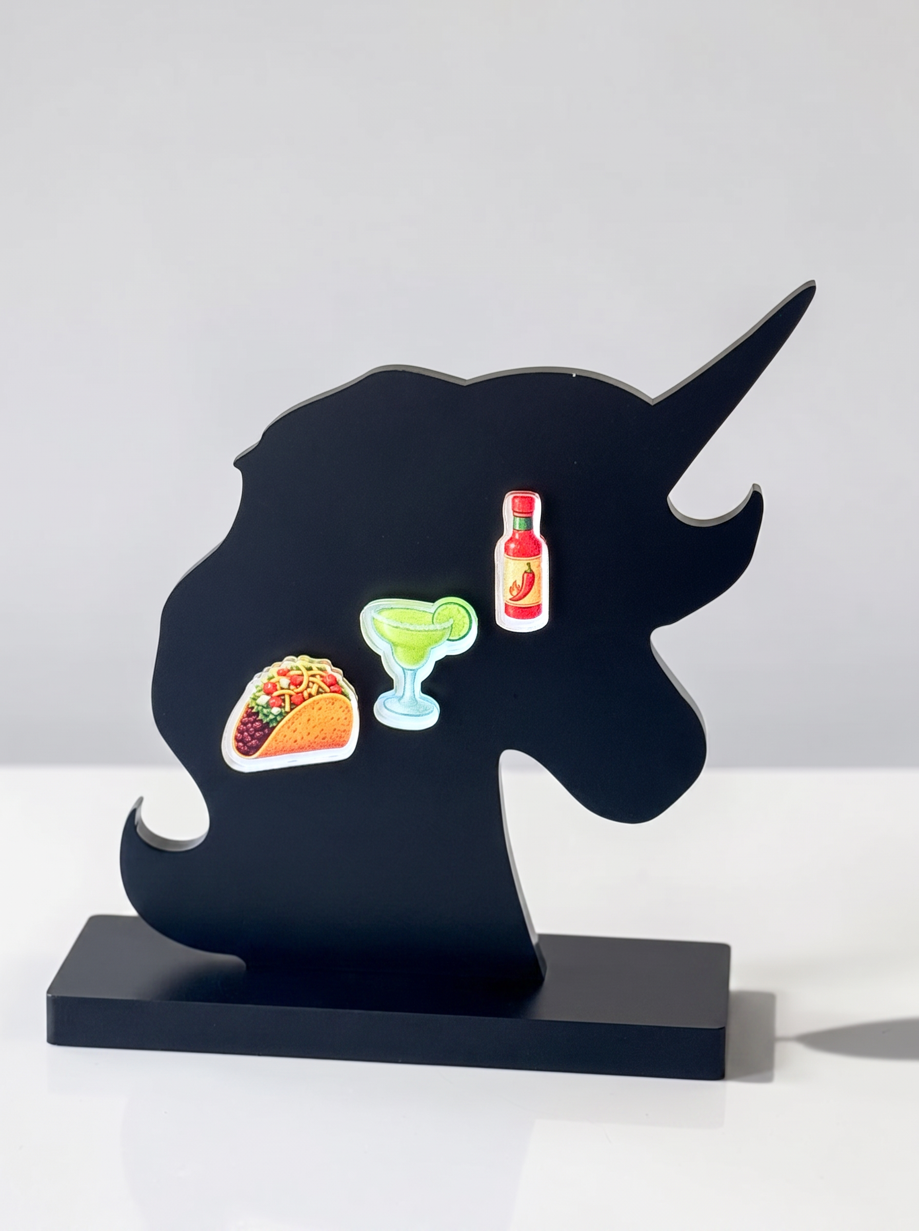 A black unicorn-shaped decorative stand with a rectangular base, featuring three small colorful food and drink earrings attached, photographed on a clean white surface against a light gray background.