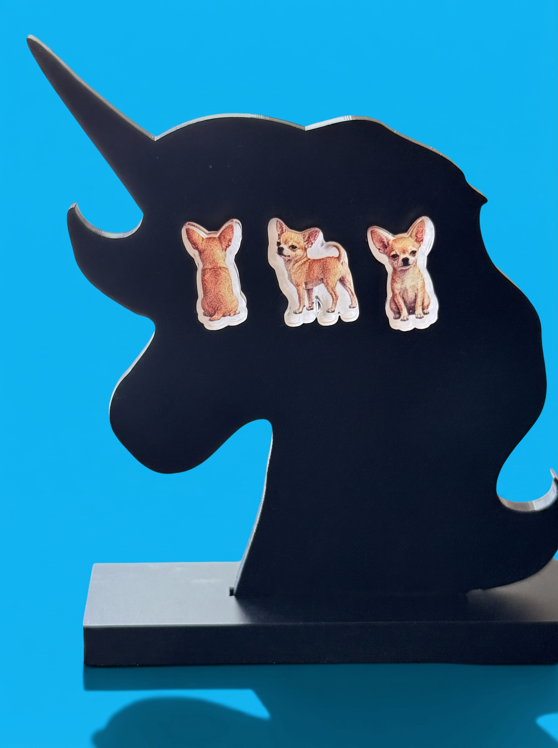 A black unicorn-shaped stand with three Chihuahua earrings set against a bright blue background.