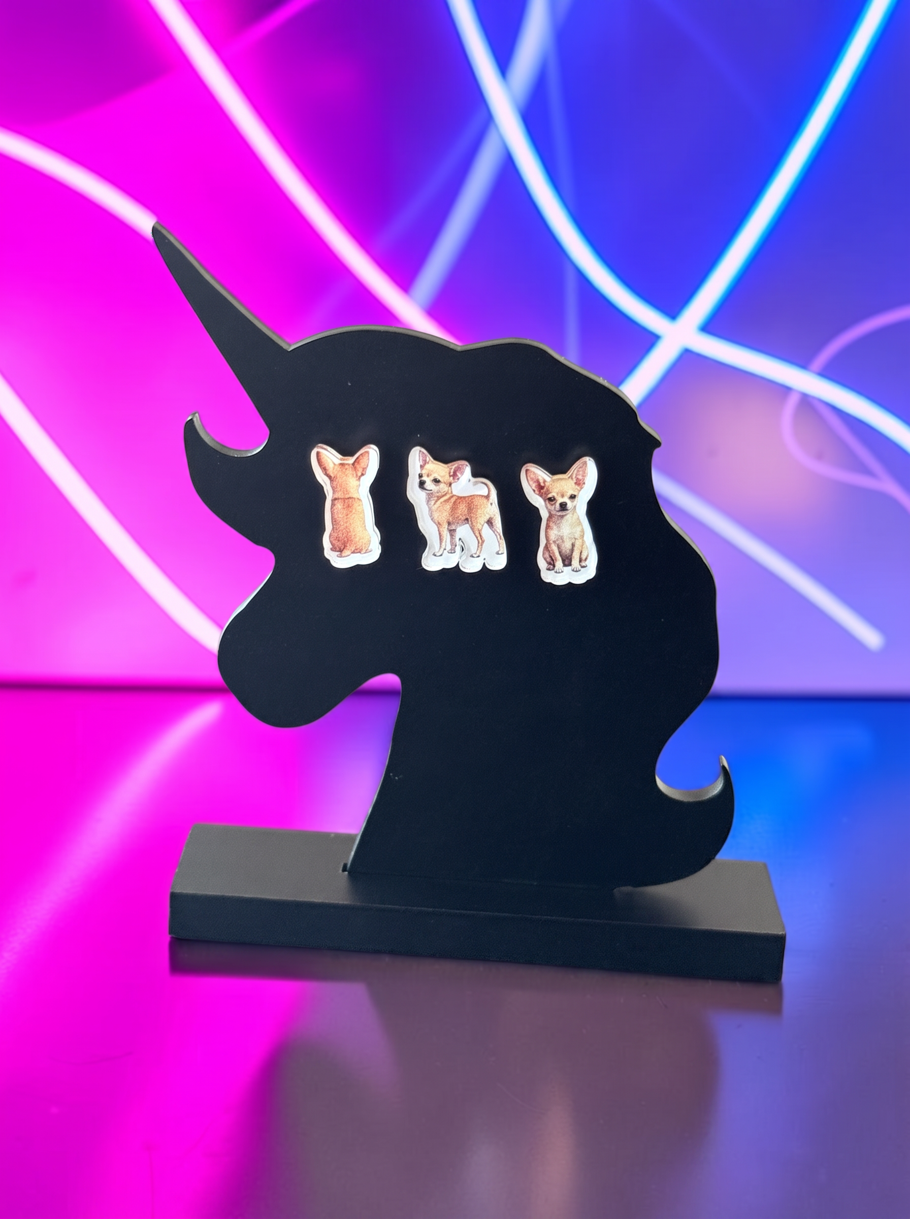 A black unicorn-shaped stand with three Chihuahua earrings set against a vibrant pink and blue neon light background.