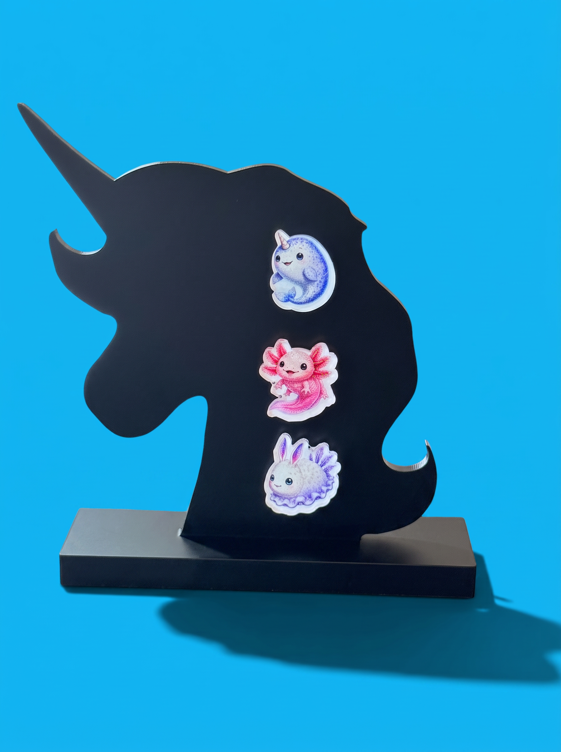A black acrylic unicorn-shaped piece with three colorful earrings. The earrings include a blue Narwhal, a pink Axolotl and a Sea Bunny which are all cute and weird at the same time. shaped stand against a bright blue background.