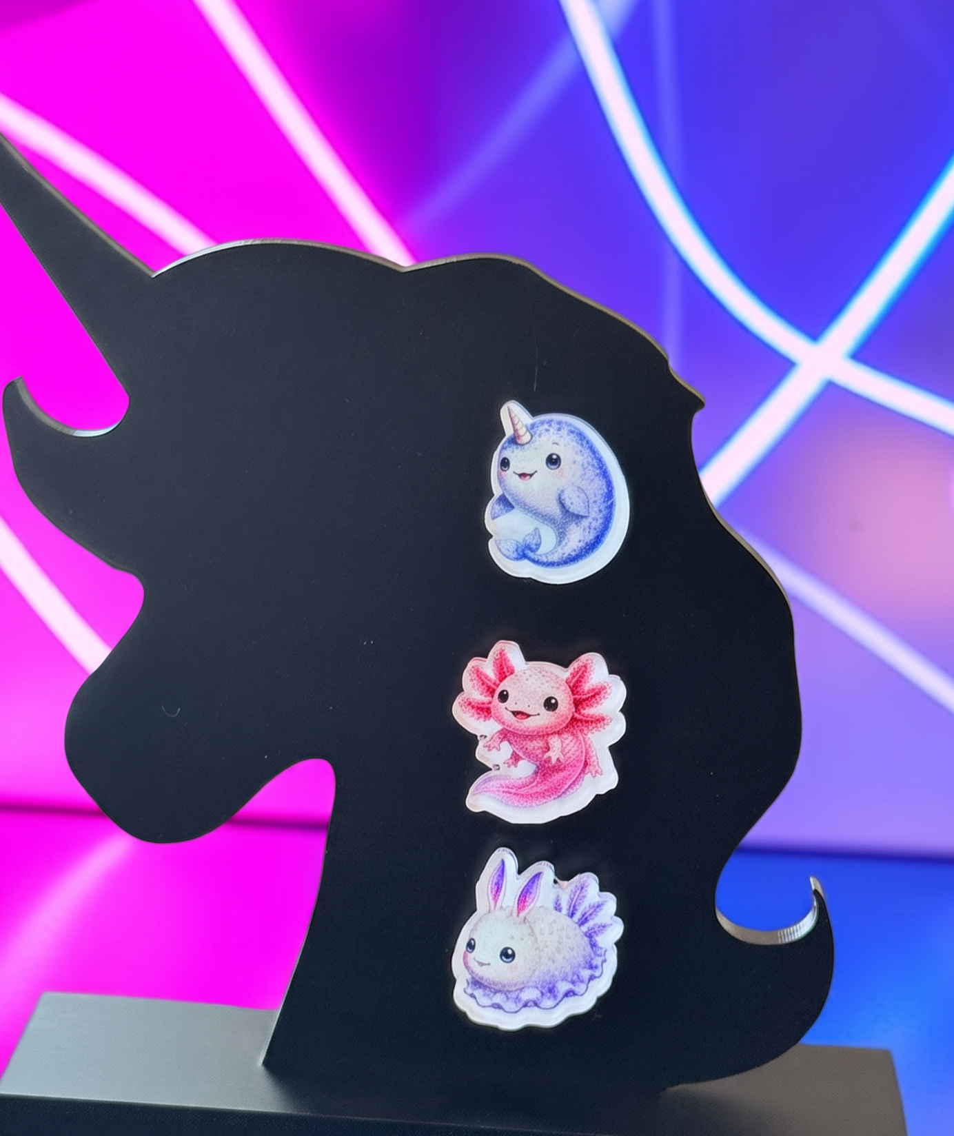 Three small illustrated  colorful earrings a blue Narwhal, a pink Axolotl and a Sea Bunny which are all cute and weird at the same time. The earrings are displayed on a black unicorn-shaped stand against a neon pink and blue background.