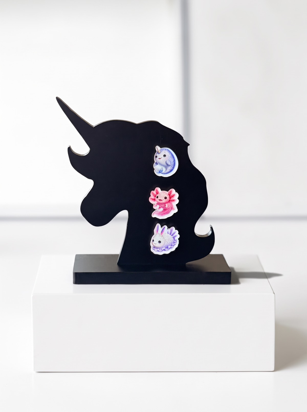 A black acryli  unicorn-shaped wall decor piece with three colorful earrings. The earrings include a blue Narwhal, a pink Axolotl and a Sea Bunny which are all cute and weird at the same time.
